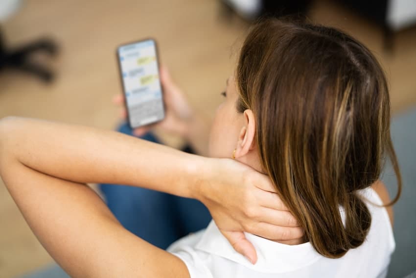 woman holding her neck in pain while looking at her phone