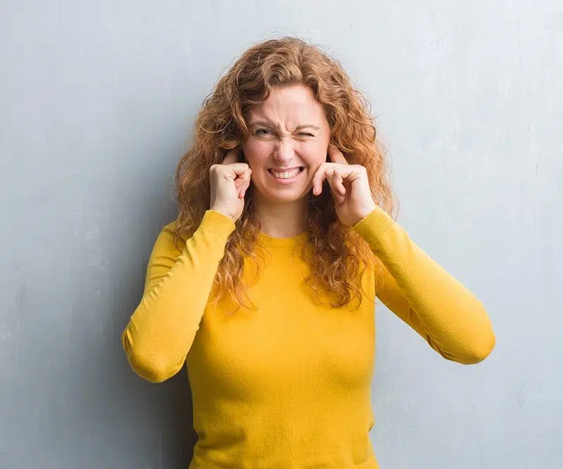 ear-pain woman in a yellow sweater plugging her ears with her fingers